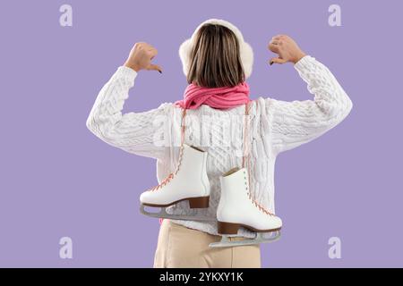 Jeune femme en vêtements d'hiver pointant vers des patins à glace sur fond lilas, vue arrière Banque D'Images