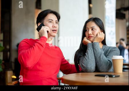 Une fille asiatique malheureuse et agacée avec une expression de mauvaise humeur est assise dans un café, se sentant ignorée par son petit ami alors qu'il parle au téléphone pendant leur c Banque D'Images