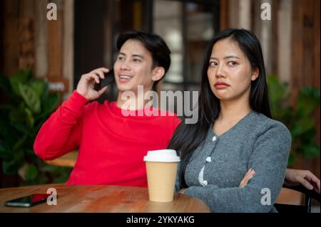 Une fille asiatique malheureuse et agacée avec une expression de mauvaise humeur est assise dans un café, se sentant ignorée par son petit ami alors qu'il parle au téléphone pendant leur c Banque D'Images