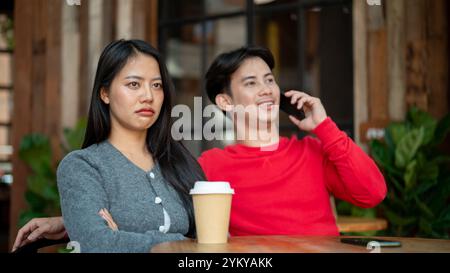 Une fille asiatique malheureuse et agacée avec une expression de mauvaise humeur est assise dans un café, se sentant ignorée par son petit ami alors qu'il parle au téléphone pendant leur c Banque D'Images