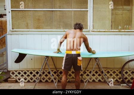 Un homme se tient devant et épile une planche de surf. Il porte un maillot de bain rayé noir et jaune Banque D'Images