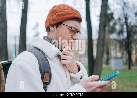 Jeune homme portant des lunettes et un bonnet orange, tenant son menton tout en lisant son smartphone dans un parc pendant une froide journée d'hiver Banque D'Images