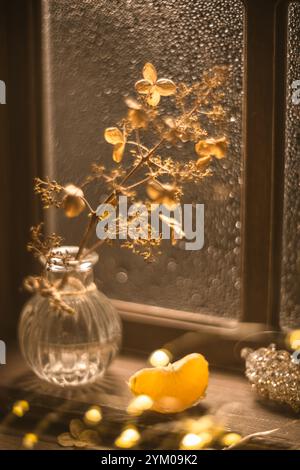 La lumière chaude du soleil illumine un vase en verre contenant des fleurs d'hortensia séchées et de la mandarine, des tranches d'orange et des lumières de Noël, créant un hiver confortable SC Banque D'Images