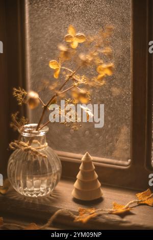 La lumière chaude du soleil illumine un vase en verre contenant des fleurs d'hortensia séchées, créant une scène automnale confortable sur un rebord de fenêtre Banque D'Images