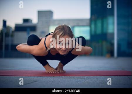 Praticien de yoga démontre la pose avancée du corbeau sur un tapis dans un cadre urbain pendant le coucher du soleil Banque D'Images