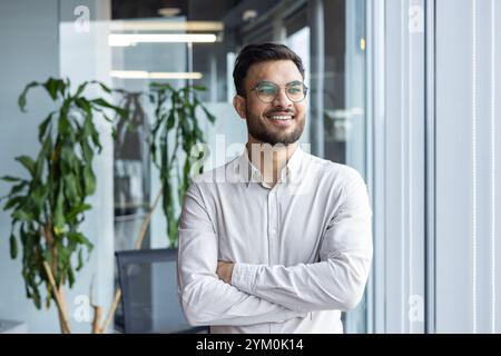 Jeune homme confiant au bureau debout près de la fenêtre, souriant et portant des lunettes. Fait preuve de positivité, de détermination et de professionnalisme dans un environnement commercial moderne. Banque D'Images