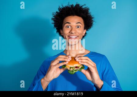 Portrait photo de jeune homme attrayant manger hamburger étonné excité habillé vêtements élégants isolés sur fond de couleur bleue Banque D'Images