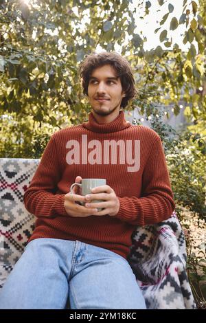 Détendez-vous dans un cadre extérieur luxuriant, un jeune et beau homme tient une tasse de vapeur tout en portant un pull élégant. La lumière du soleil filtre à travers les feuilles Banque D'Images