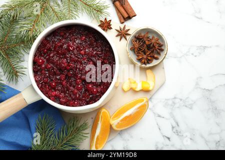 Savoureuse sauce aux canneberges dans une casserole, épices, branches d'orange et de sapin sur table en marbre blanc, pose à plat. Espace pour le texte Banque D'Images