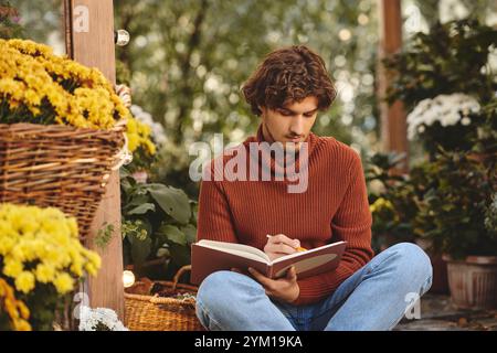 Un beau jeune homme est assis sur le sol dans un jardin rempli de fleurs colorées, profondément concentré sur l'écriture dans un cahier. L'atmosphère réconfortante o Banque D'Images