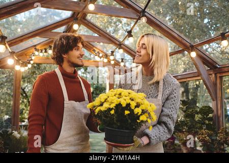 Serre bien éclairée remplie de plantes vibrantes. Un jeune couple beau s'engage dans une conversation joyeuse tout en admirant un pot de fl jaune joyeux Banque D'Images