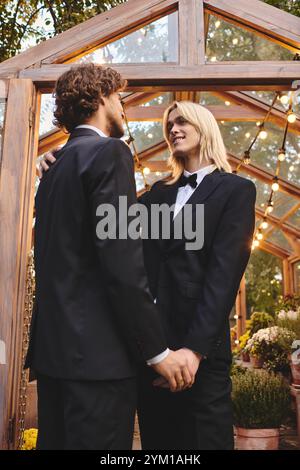 Deux hommes élégants, vêtus de smoking assortis, se tiennent intimement dans une charmante serre. Banque D'Images