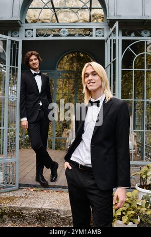 Deux jeunes hommes élégants vêtus de costumes élégants posent dans un jardin enchanteur. L'un se tient détendu tandis que l'autre sourit en toute confiance, capturant un moment Banque D'Images