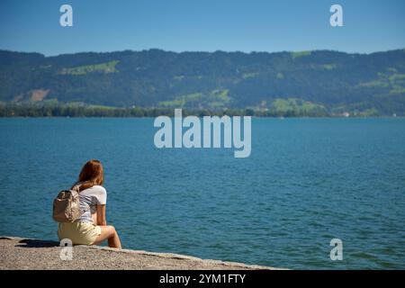 Une jeune femme profite d'un moment de sérénité au bord d'un lac entouré de montagnes et de verdure luxuriante par une journée ensoleillée. Blog de voyage. Aventure, concept de vacances. Ser Banque D'Images