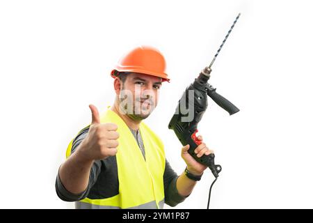 Homme de construction adulte amical dans le gilet fluorescent et le casque de sécurité orange tenant l'outil de perceuse électrique faisant pouce vers le haut comme geste isolé sur le bac blanc Banque D'Images