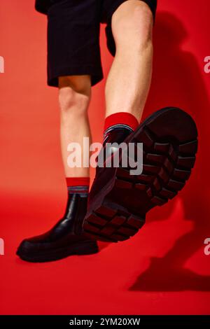 Un homme met en valeur la mode tendance des fêtes avec des bottes noires élégantes, des chaussettes rouges éclatantes et une pose confiante sur un fond rouge saisissant. Ce styl Banque D'Images