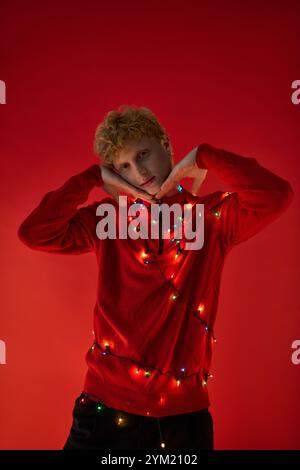 Un homme pose de manière ludique sur un fond rouge audacieux, drapé de lumières de Noël colorées. Son pull rouge festif capture l'esprit de la mer de vacances Banque D'Images