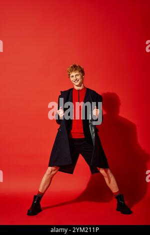 Un homme élégant met en valeur la mode de vacances avec confiance. Vêtu d'un haut rouge vif et d'un manteau élégant, il se tient sur un fond rouge saisissant, e. Banque D'Images