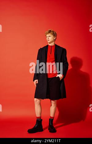 Un homme élégant présente la mode des fêtes avec un haut rouge confortable et un élégant manteau noir. Il se tient confiant sur un fond rouge vif, évoquant un Banque D'Images