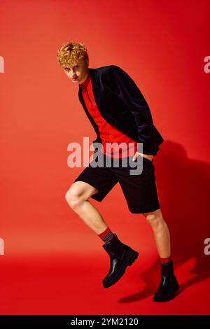 Un homme élégant présente la dernière mode des fêtes sur fond rouge vif. Il arbore une tenue tendance avec un short et une veste élégante, perf Banque D'Images