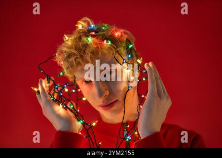 Un jeune homme élégant montre sa créativité en enveloppant des lumières colorées des fêtes autour de sa tête. Banque D'Images