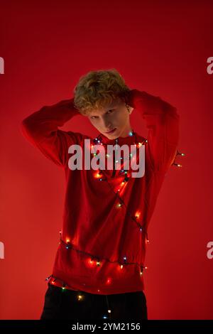 Un jeune homme présente sa tenue de vacances élégante ornée de lumières de Noël colorées sur un fond rouge vif. Cet arrangement festif captu Banque D'Images