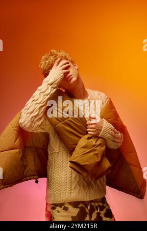 Un jeune homme met en valeur la mode des fêtes à la mode, enfilant un pull confortable et tenant une veste élégante, entouré de couleurs festives vibrantes. Le réglage e Banque D'Images