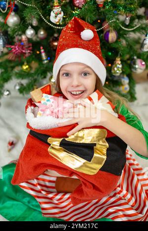 Une fille en tenue festive s'accroche joyeusement plusieurs cadeaux enveloppés colorés près d'un arbre de Noël orné d'ornements. Banque D'Images