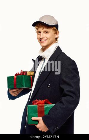 Arborant un manteau d'hiver tendance et une casquette, un jeune homme poutres tout en tenant deux joliment emballés cadeaux de Noël. Banque D'Images