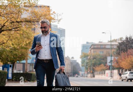 Un bel homme d'affaires mature utilisant l'application de téléphone mobile SMS tout en marchant dans la rue de la ville. Banque D'Images