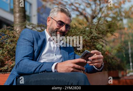 Un bel homme d'affaires heureux utilisant l'application de téléphone mobile SMS tout en étant assis sur le banc du parc. Banque D'Images