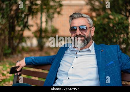 Un bel homme d'affaires mûr heureux se détache tout en étant assis sur le banc du parc. Banque D'Images