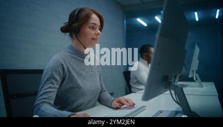 Bureau du centre d'appels la nuit : spécialiste sérieuse de l'assistance technique féminine dans les micro-casques/oreillettes surveille les données sur PC, fournit un service d'assistance client en ligne par appel. Équipe d'opérateurs de hotline multiethniques travaillant. Banque D'Images