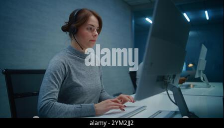 Bureau du centre d'appels la nuit : spécialiste sérieuse de l'assistance technique féminine dans les micro-casques/oreillettes surveille les données sur PC, fournit un service d'assistance client en ligne par appel. Équipe d'opérateurs de hotline multiethniques travaillant. Banque D'Images