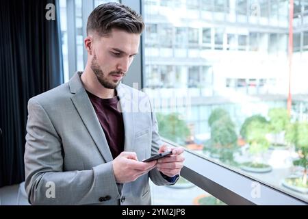 17 novembre 2024 : un jeune homme en costume gris se tient debout dans un bureau moderne, tapant intensément sur son smartphone *** Ein Junger Mann in einem grauen Anzug steht in einem modernen Büro und tippt konzentriert auf seinem smartphone Banque D'Images