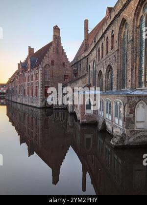 Des bâtiments médiévaux en briques se reflètent dans un canal tranquille au coucher du soleil, créant une atmosphère sereine. Bruges, Belgique, Europe Banque D'Images