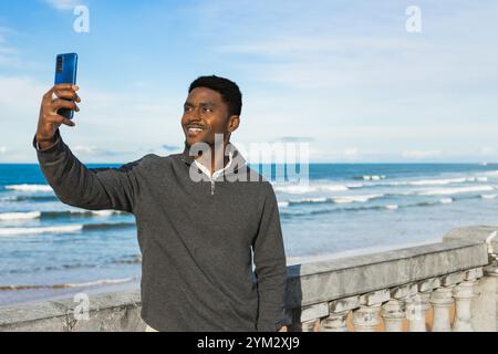 Smiling Man prend un selfie par l'océan par une journée ensoleillée, capturant le ciel bleu et les vagues en arrière-plan. Il se tient près d'une balustrade en pierre, portant un Banque D'Images