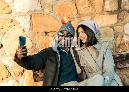Couple souriant portant des vêtements d'hiver confortables prend un selfie contre un mur de pierre à l'extérieur. ils sont habillés chaudement avec des vestes, des chapeaux et des lunettes de soleil, Banque D'Images