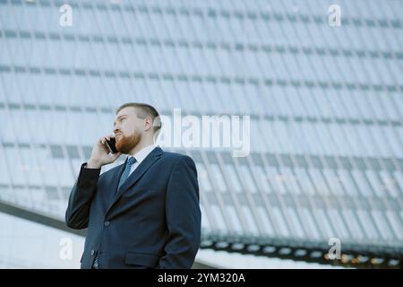 Homme d'affaires portant costume, parlant sur téléphone portable, debout à l'extérieur de l'immeuble de bureaux en verre contemporain, se concentrant sur la conversation Banque D'Images