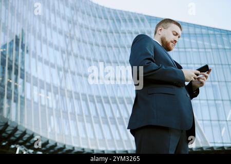 Homme d'affaires en tenue formelle examinant des documents tout en se tenant debout à l'extérieur du bâtiment de verre moderne pendant la journée. Structure haute en arrière-plan reflétant le ciel clair Banque D'Images