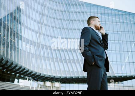 Personne portant le costume d'affaires parlant sur le téléphone portable à l'extérieur du bâtiment en verre moderne, montrant l'accent sur le travail professionnel dans l'environnement urbain Banque D'Images