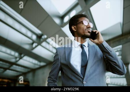 Homme d'affaires en costume formel parlant sur le téléphone portable tout en se tenant sous les puits de lumière modernes dans le centre d'affaires. Expression confiante et comportement professionnel capturés Banque D'Images