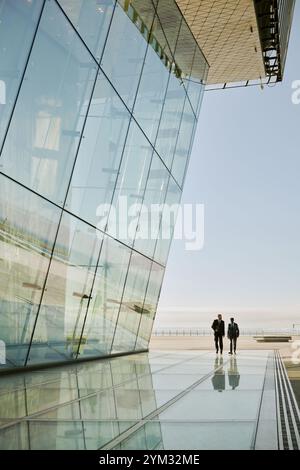 Façade en verre réfléchissant d'un bâtiment moderne projetant des motifs élégants sur le paysage urbain environnant montrant deux individus marchant le long de la surface miroir à la lumière du soleil Banque D'Images