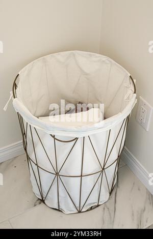 Un panier blanc avec un chiffon blanc est assis sur un sol en carreaux blancs. Le panier est vide et a une conception en treillis métallique Banque D'Images