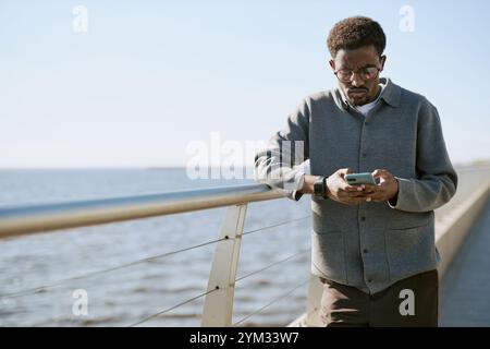 Jeune homme afro-américain utilisant un smartphone tout en se tenant près d'une rampe de front de mer par une journée brillante. Expression focalisée sur le visage de l'homme tout en naviguant sur son appareil Banque D'Images