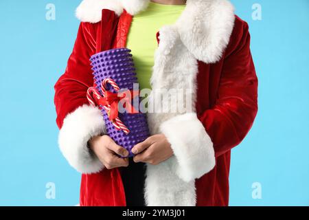 Jeune homme sportif en costume de Père Noël avec des cannes de bonbons de Noël et rouleau de mousse sur fond bleu, gros plan Banque D'Images