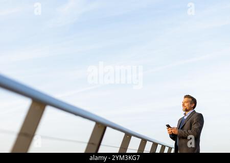 Vêtu d'un costume formel, un homme d'affaires se tient debout sur un pont élégant, concentré sur son smartphone au coucher du soleil. L'horizon de la ville crée un arrière-plan magnifique Banque D'Images