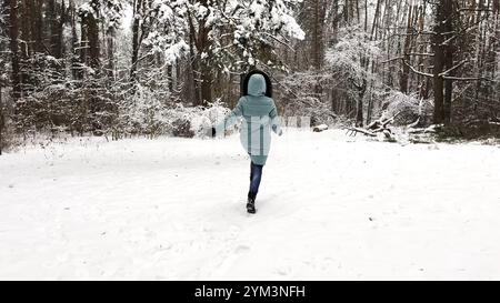 Jeune fille courant sur la piste à travers la forêt enneigée. Femme heureuse faisant du jogging sur le chemin à Woodland. Dame joyeuse s'amusant et profitant de l'hiver. Concept Banque D'Images