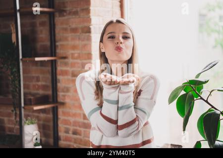 Jeune femme souffle un baiser ludique à la maison, profitant d'un week-end de détente dans un salon confortable avec la lumière naturelle Banque D'Images