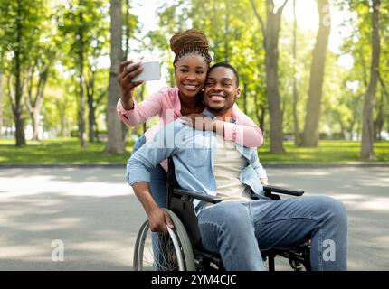 Une femme noire heureuse et son petit ami handicapé en fauteuil roulant emportant le selfie ensemble, se tenant dans le parc de la ville Banque D'Images